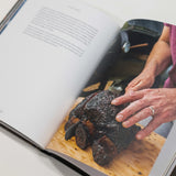 An open cookbook or recipe book lying on a light surface, showing a printed photo of hands handling a dark cooked meat on a wooden board on one page.