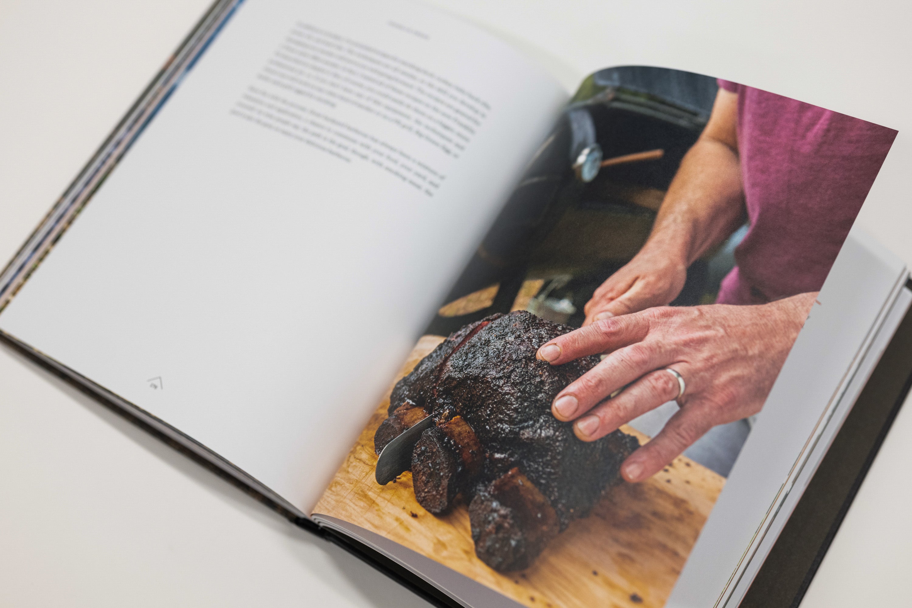 An open cookbook or recipe book lying on a light surface, showing a printed photo of hands handling a dark cooked meat on a wooden board on one page.