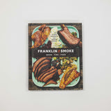The cookbook 'Franklin Smoke: Wood Fire Food' by Aaron Franklin and Jordan McCullough, featuring a rustic cover with a photo of grilled meats and a black banner with the title, displayed on a light gray background.