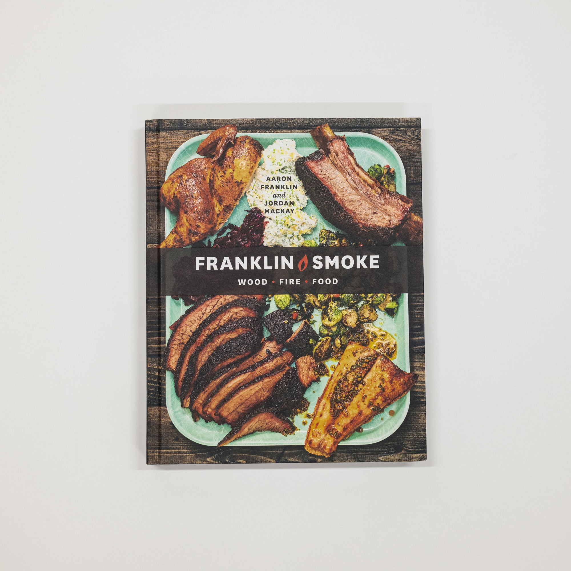 The cookbook 'Franklin Smoke: Wood Fire Food' by Aaron Franklin and Jordan McCullough, featuring a rustic cover with a photo of grilled meats and a black banner with the title, displayed on a light gray background.