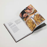 An open hardcover cookbook photographed on a clean white background. The right page features a collage of food photography, including hands preparing food and plated dishes, while the left page contains printed text.