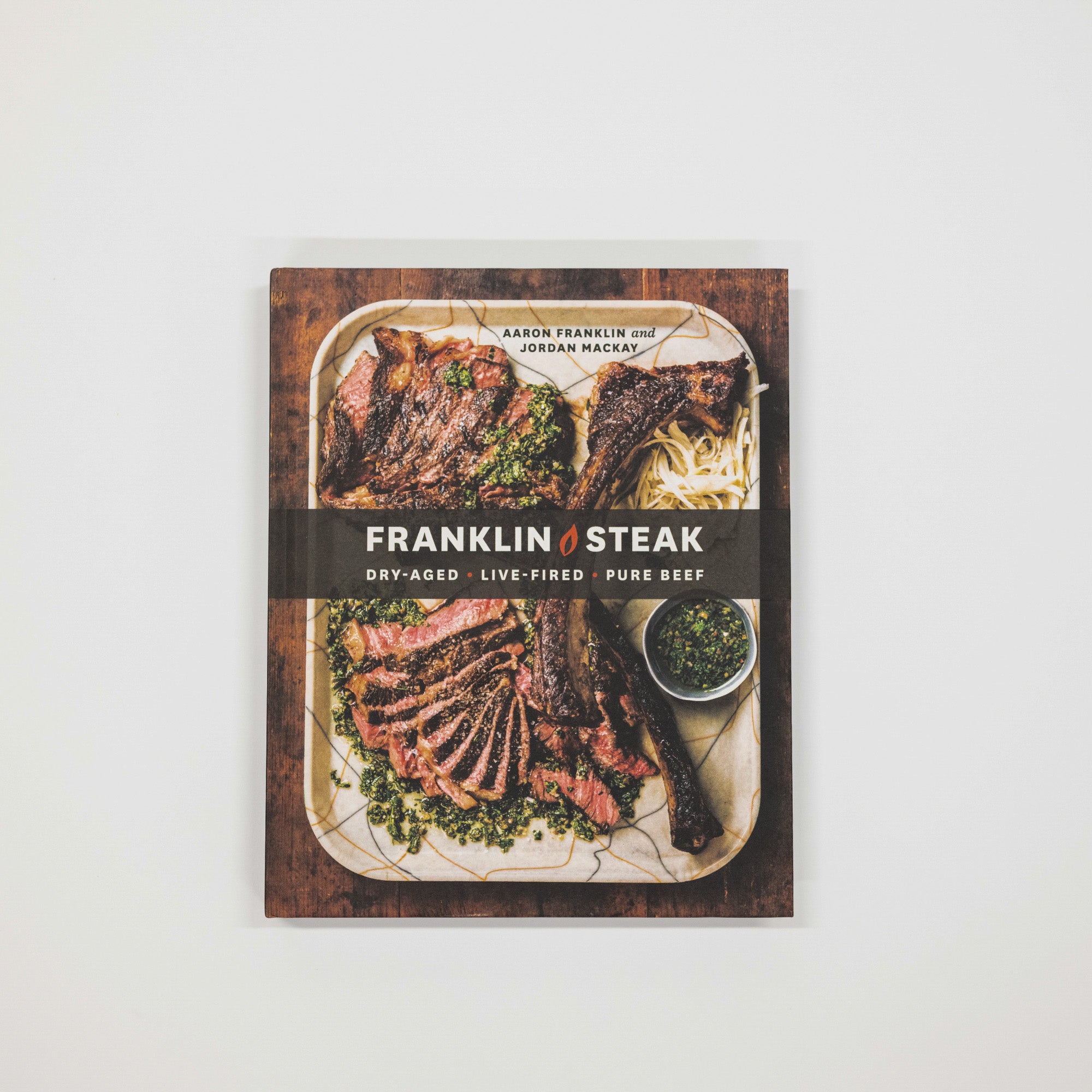 The book cover of 'Franklin Steak' by Aaron Franklin and Jordan Macask is shown centered against a plain white background. The cover features a professional food photograph of sliced steak on a serving platter with green herbs and fries, overlaid with a dark brown banner containing the white title text.