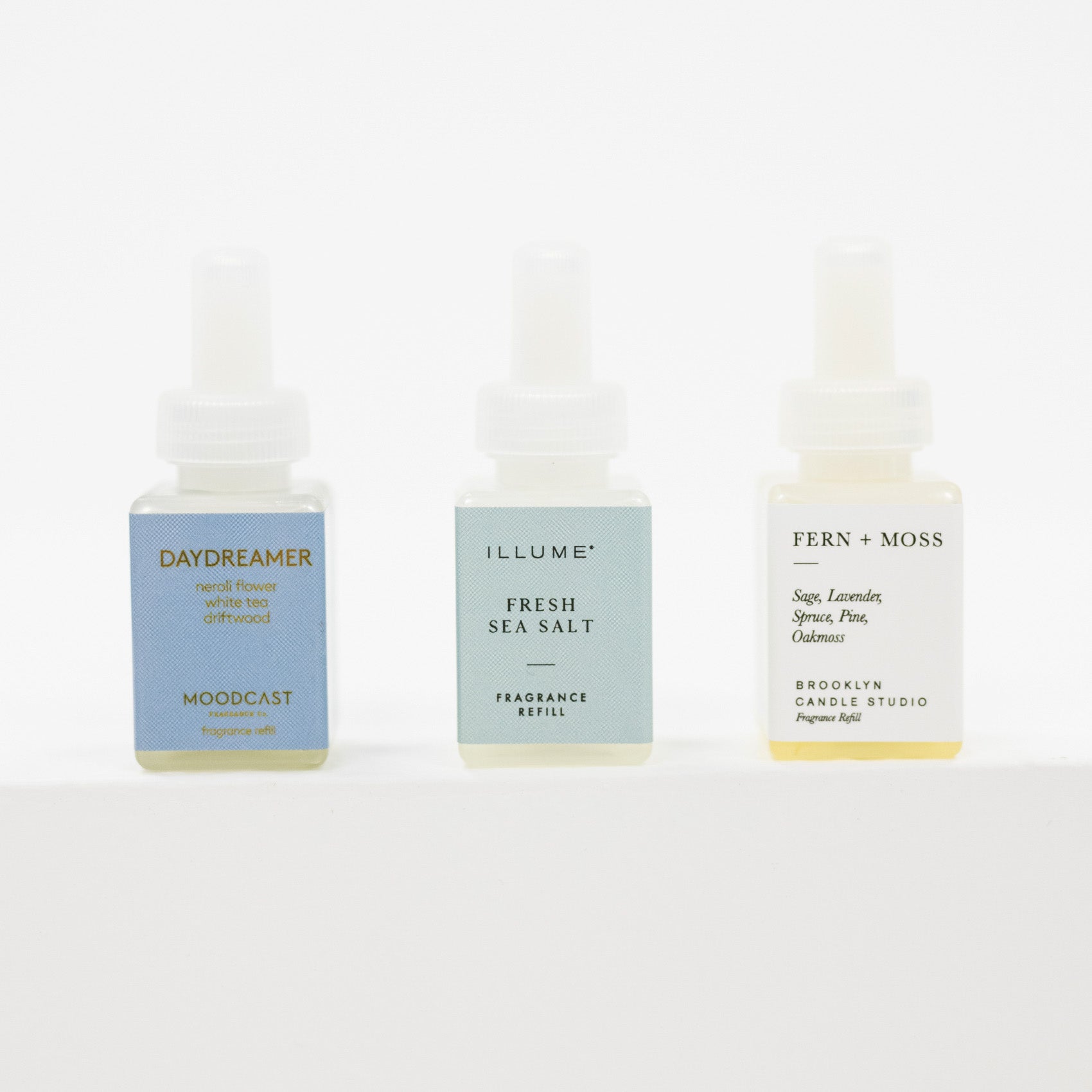 Three small pura fragrance refill bottles on a clean white background: Daydreamer by Moodcast with a blue label, Illuminate Fresh Sea Salt with a pale blue label, and Fern + Moss by Brooklyn Candle Studio with a white label.