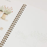 An open spiral-bound art journal or sketchbook showing a watercolor page with potted plants on the left and a lightly sketched plant illustration on the right.