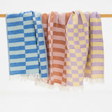 Three woven kitchen towels draped over a wooden rod against a white background, featuring blue and light blue checkered patterns, terracotta and pink checkered patterns, lavender and yellow checkered patterns, and white fringed edges.