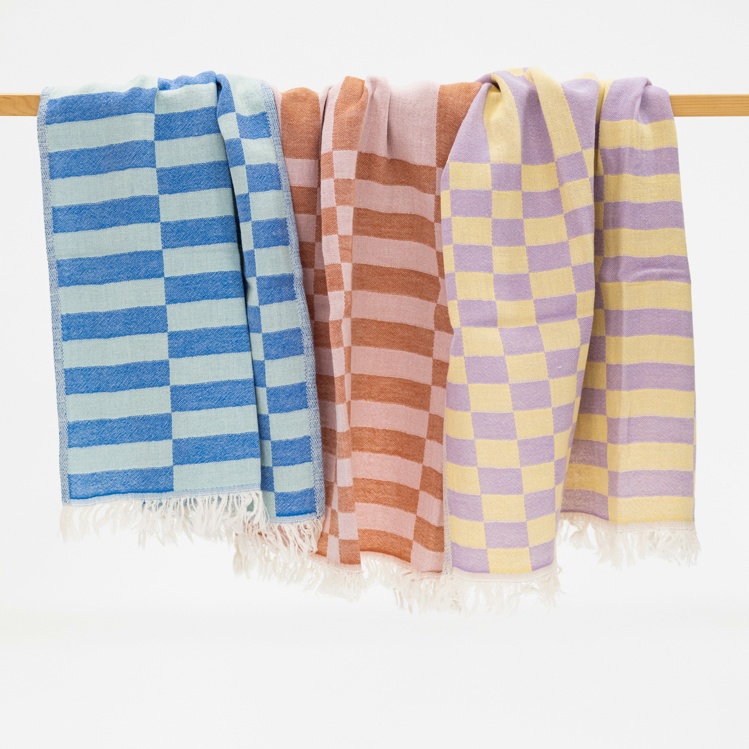 Three woven kitchen towels draped over a wooden rod against a white background, featuring blue and light blue checkered patterns, terracotta and pink checkered patterns, lavender and yellow checkered patterns, and white fringed edges.