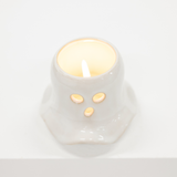 A small white ceramic candle holder shaped like a cute animal face with two circular eyes and a triangular nose, shown lit with a warm yellow glow against a clean white background.