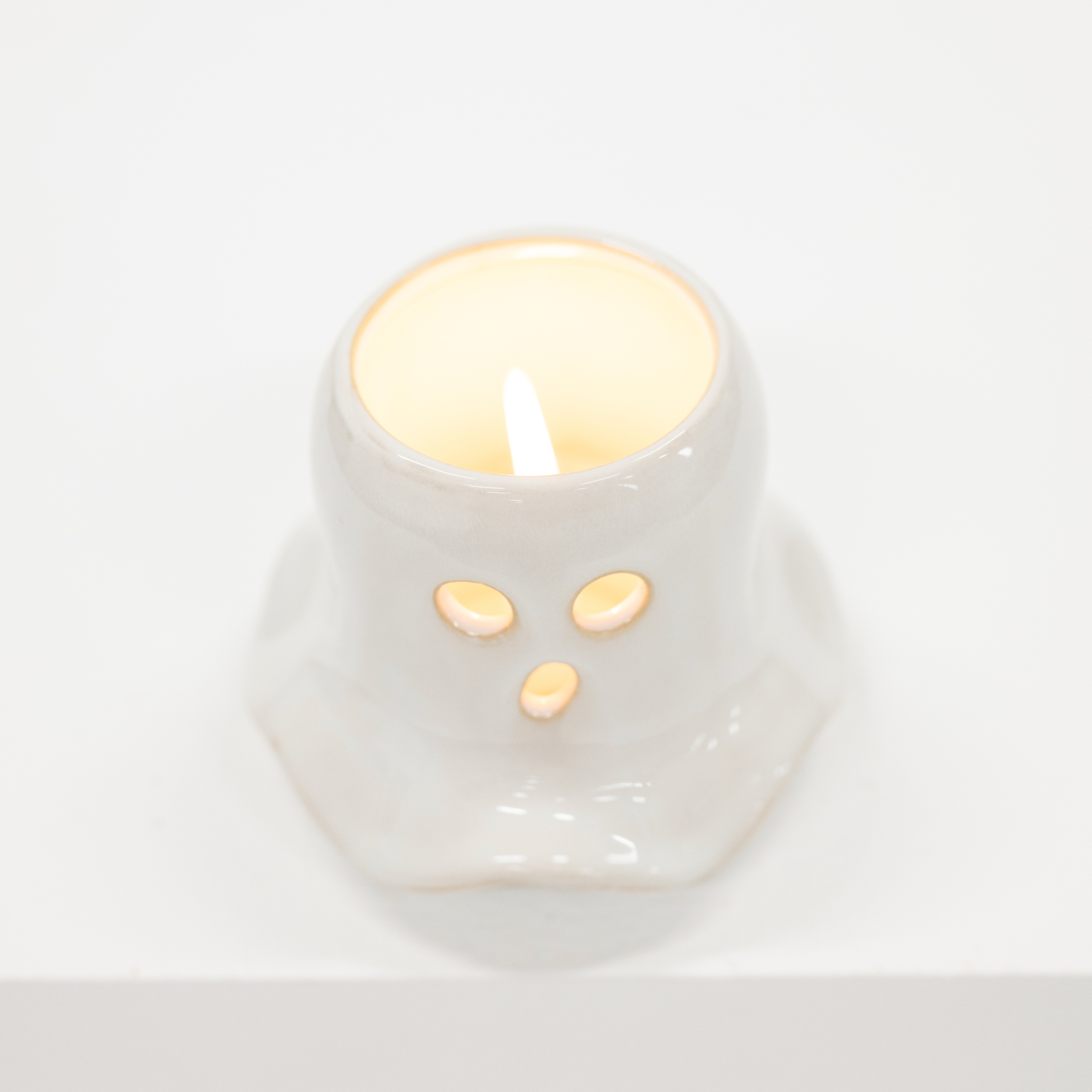 A small white ceramic candle holder shaped like a cute animal face with two circular eyes and a triangular nose, shown lit with a warm yellow glow against a clean white background.