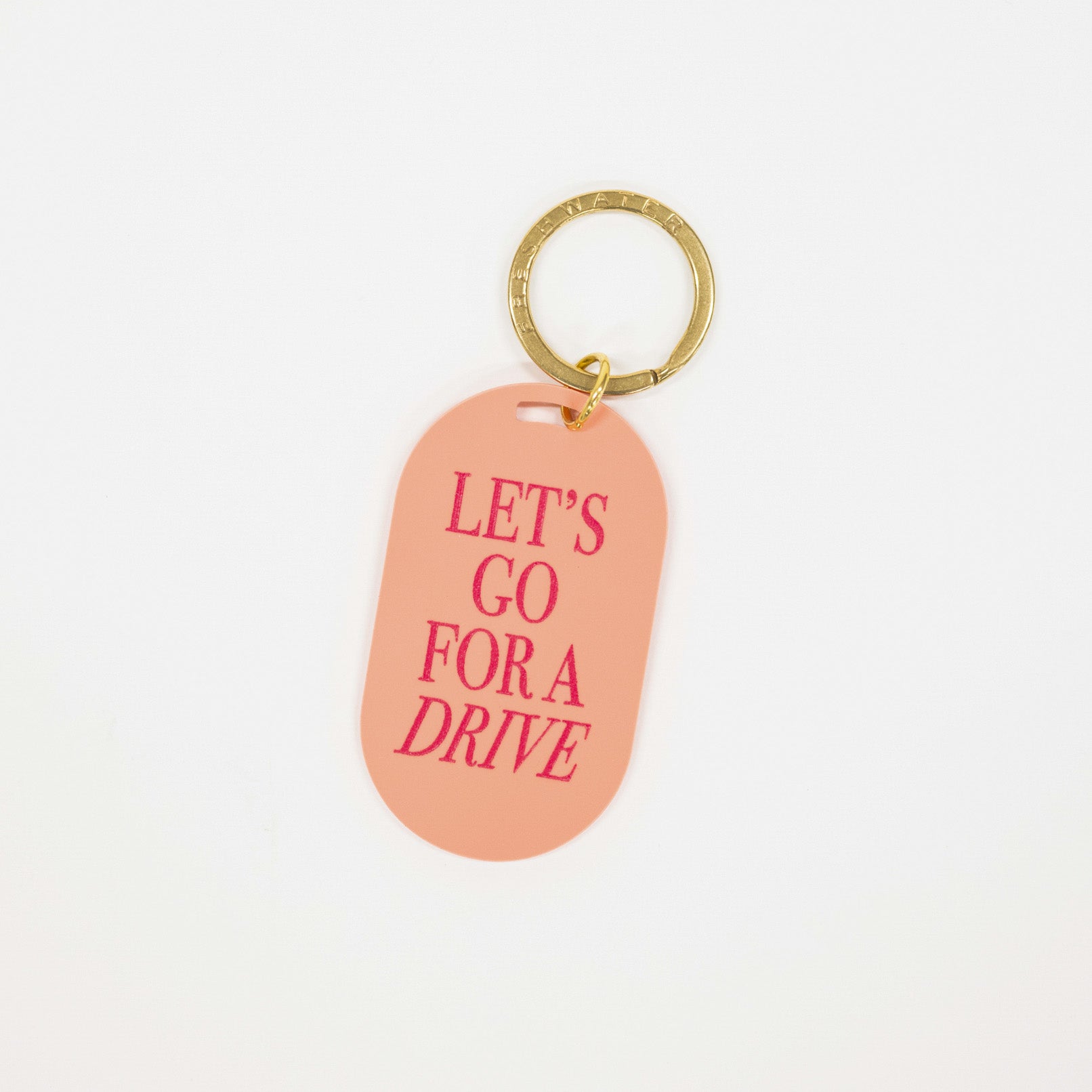 A peach-colored dog tag keychain with the text 'LET'S GO FOR A DRIVE' in pink, attached to a gold split ring, shown on a plain white background.