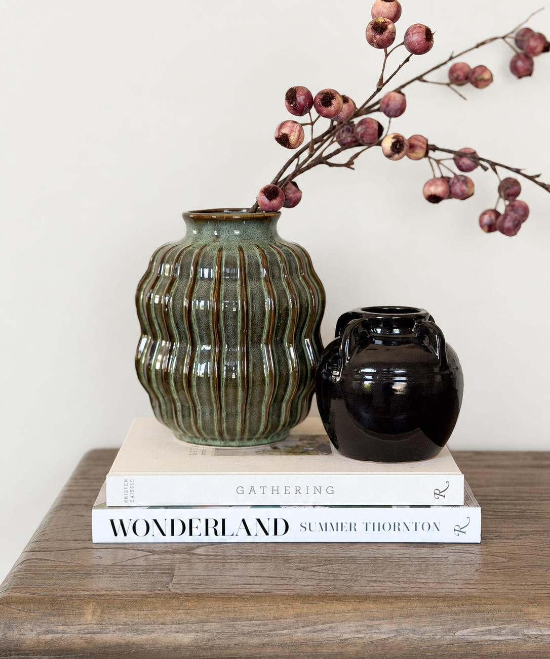 a green ceramic vase styled with a twig of faux berries, a small black vase and two books on a wooden table