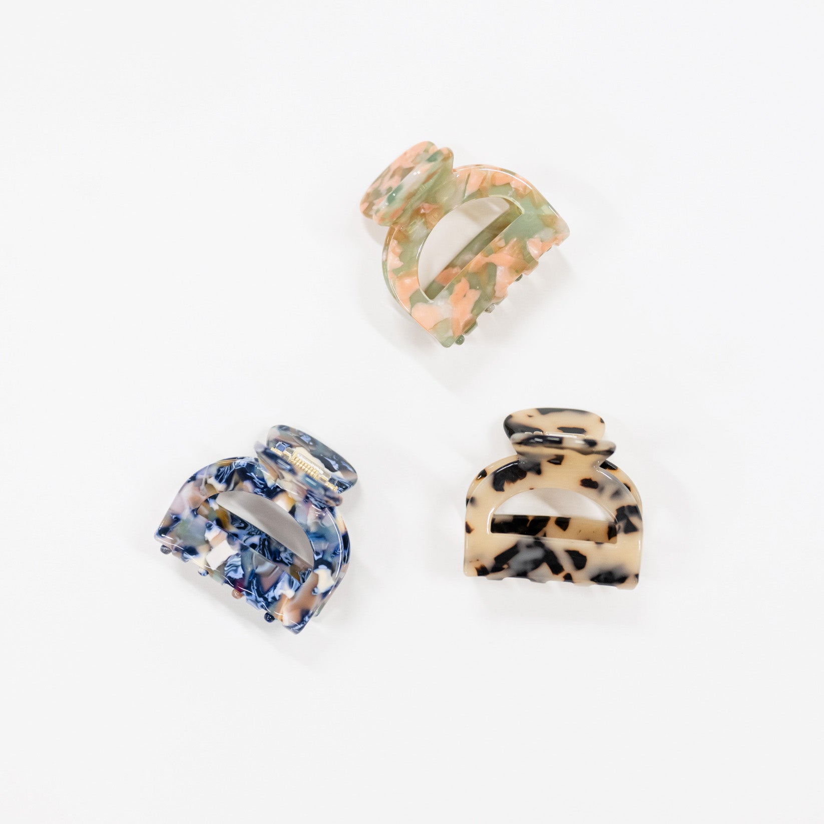 Three tortoiseshell-style hair claw clips in different color patterns (blue and white marbled, peach and green marbled, and beige and black spotted) arranged on a plain white background.