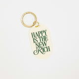 cream colored acrylic oval shaped keychain with the phrase "happy is the new rich" written in forest green. on a gold metal keychain loop