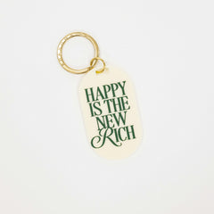 cream colored acrylic oval shaped keychain with the phrase "happy is the new rich" written in forest green. on a gold metal keychain loop
