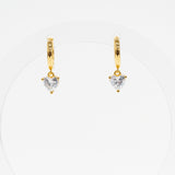 A pair of gold huggie hoop earrings with a small heart-shaped clear gemstone dangling from the center, displayed on a clean white background.