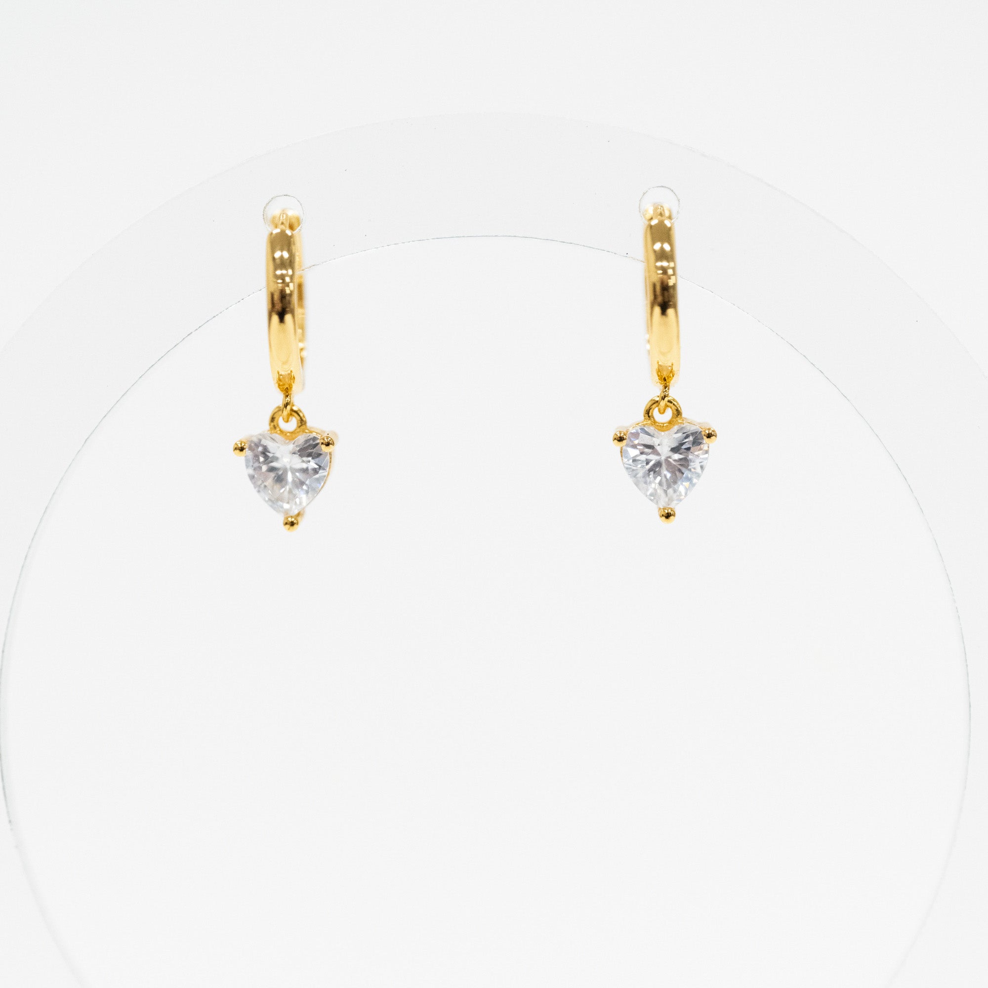 A pair of gold huggie hoop earrings with a small heart-shaped clear gemstone dangling from the center, displayed on a clean white background.