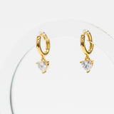 A pair of gold huggie hoop earrings with dangling heart-shaped clear gemstones, displayed on a clear acrylic stand against a white background.