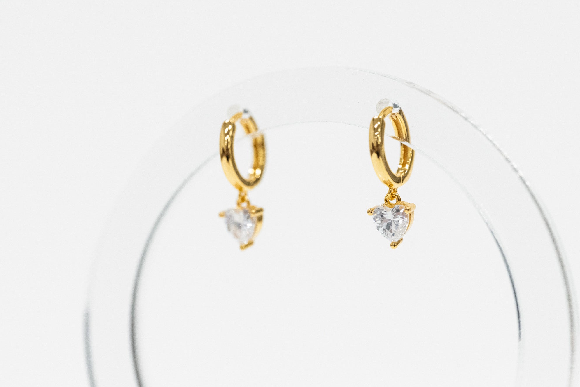 A pair of gold huggie hoop earrings with dangling heart-shaped clear gemstones, displayed on a clear acrylic stand against a white background.