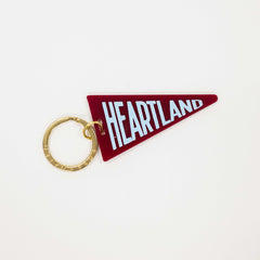 A heart-shaped red keychain featuring the word 'HEARTLAND' in white block letters, attached to a gold split key ring, shown on a plain white background.