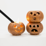 Three small jack-o'-lantern-shaped candle holders with carved faces, one lit and being held by a black lighter, with two more stacked on the right against a clean white background.
