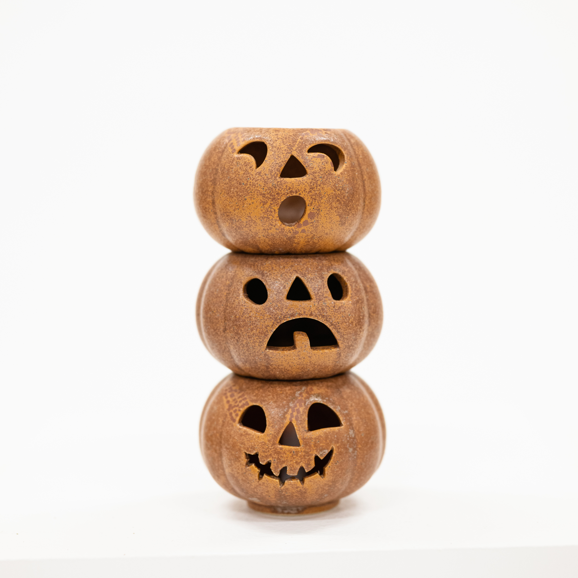 Three small carved jack-o'-lantern pumpkins stacked vertically, made of textured brown material with cutout facial features, on a plain white background.