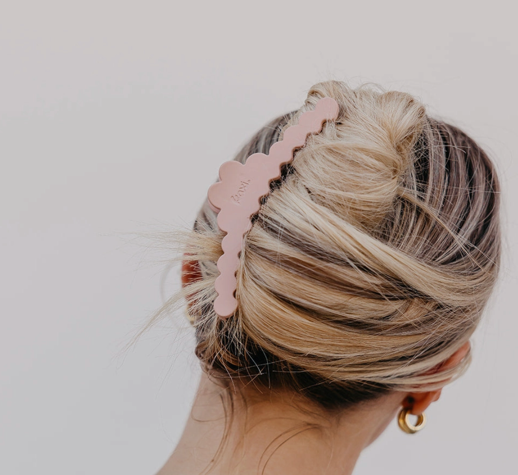 A blonde woman shown from the back wearing a dusty pink scalloped claw hair clip holding her hair in a low bun, against a plain light gray background.