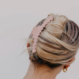 A blonde woman shown from the back wearing a dusty pink scalloped claw hair clip holding her hair in a low bun, against a plain light gray background.