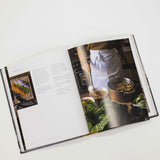An open hardcover cookbook laid flat on a light gray background, showing printed text on the left page and a large food photography page on the right with a person in a kitchen preparing food.