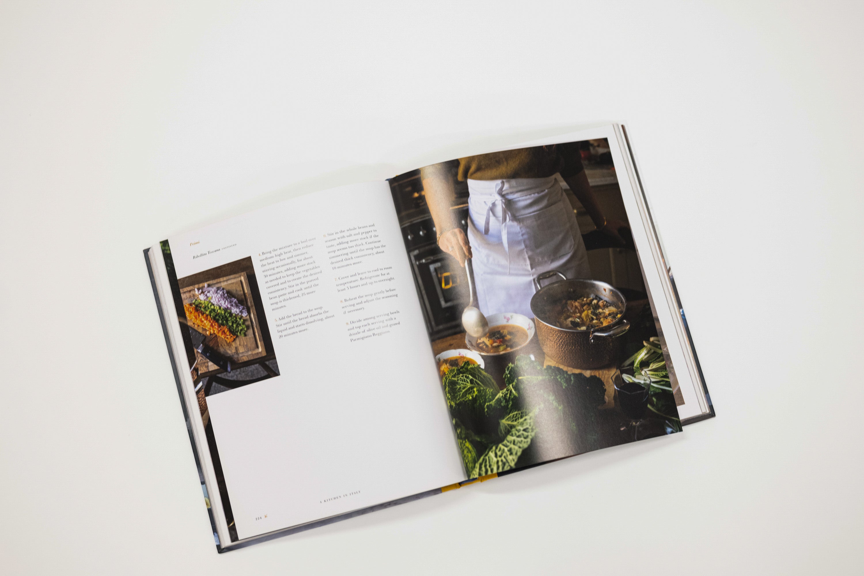 An open hardcover cookbook laid flat on a light gray background, showing printed text on the left page and a large food photography page on the right with a person in a kitchen preparing food.