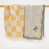 Two woven kitchen towels hanging from a wooden rod against a white background. The left towel has a yellow and cream checkerboard pattern with white fringe.