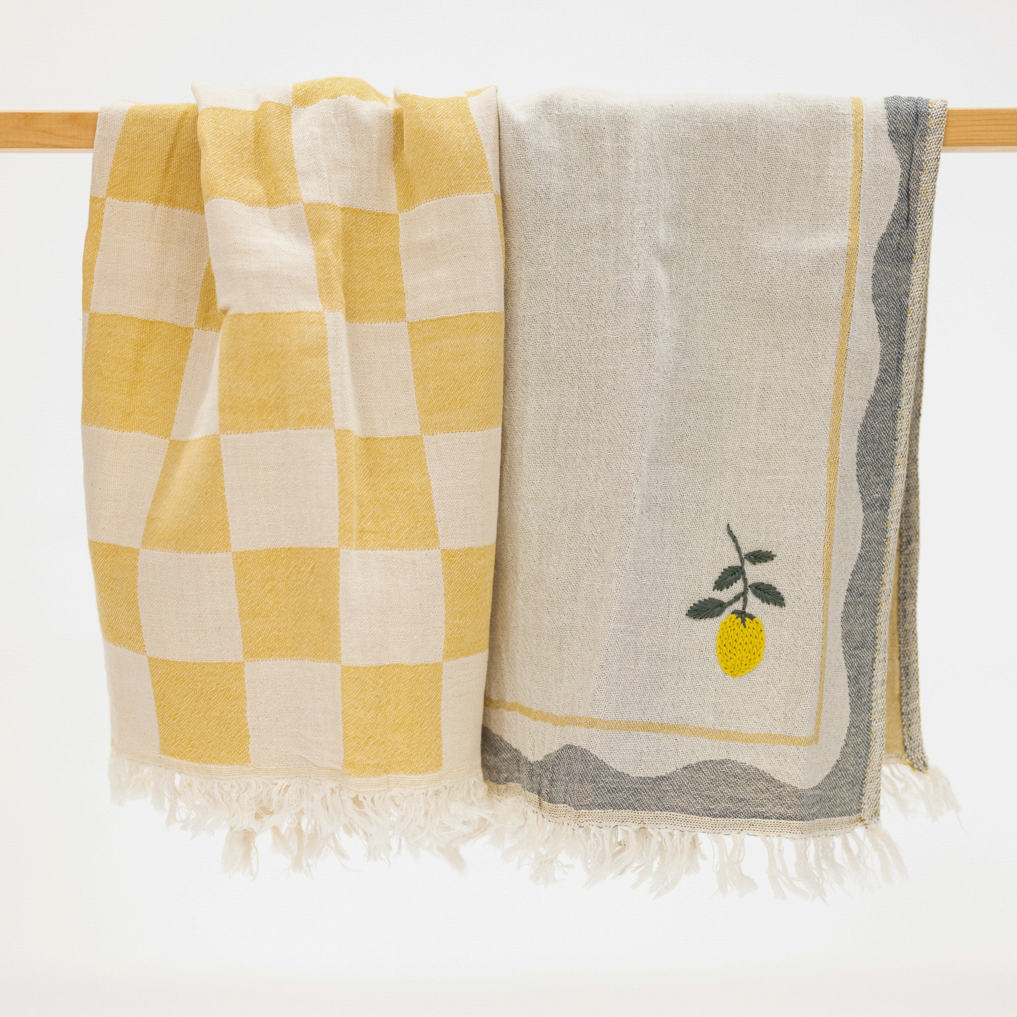 Two woven kitchen towels hanging from a wooden rod against a white background. The left towel has a yellow and cream checkerboard pattern with white fringe.