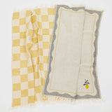 Two woven kitchen towels with fringed edges laid flat on a white background. One has a yellow and cream checkerboard pattern, and the other has a gray border with a small yellow lemon and green stem motif on the bottom right.