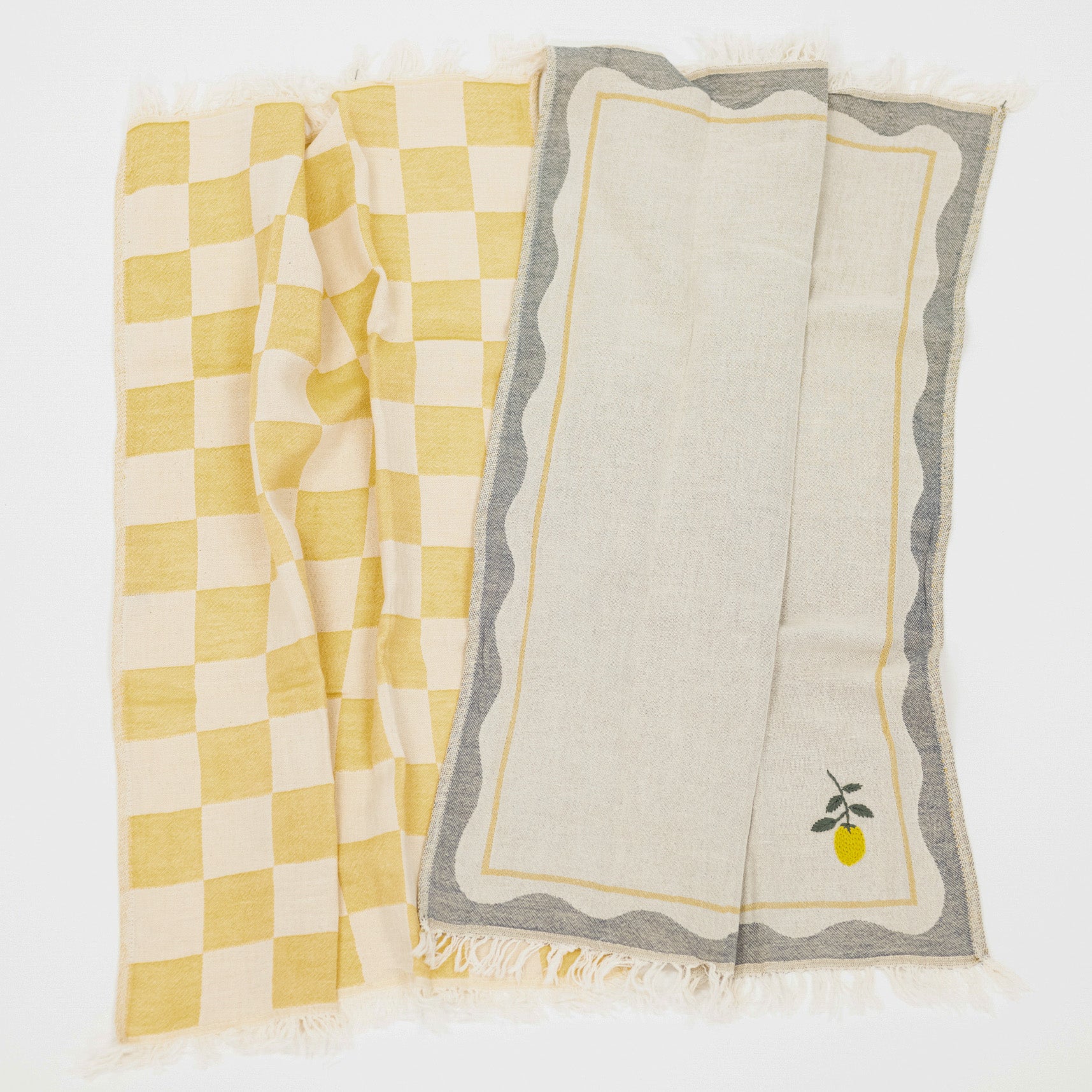 Two woven kitchen towels with fringed edges laid flat on a white background. One has a yellow and cream checkerboard pattern, and the other has a gray border with a small yellow lemon and green stem motif on the bottom right.