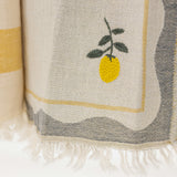 A close-up of a woven textile with fringe, featuring a yellow lemon embroidery on a cream background with gray and yellow border details.