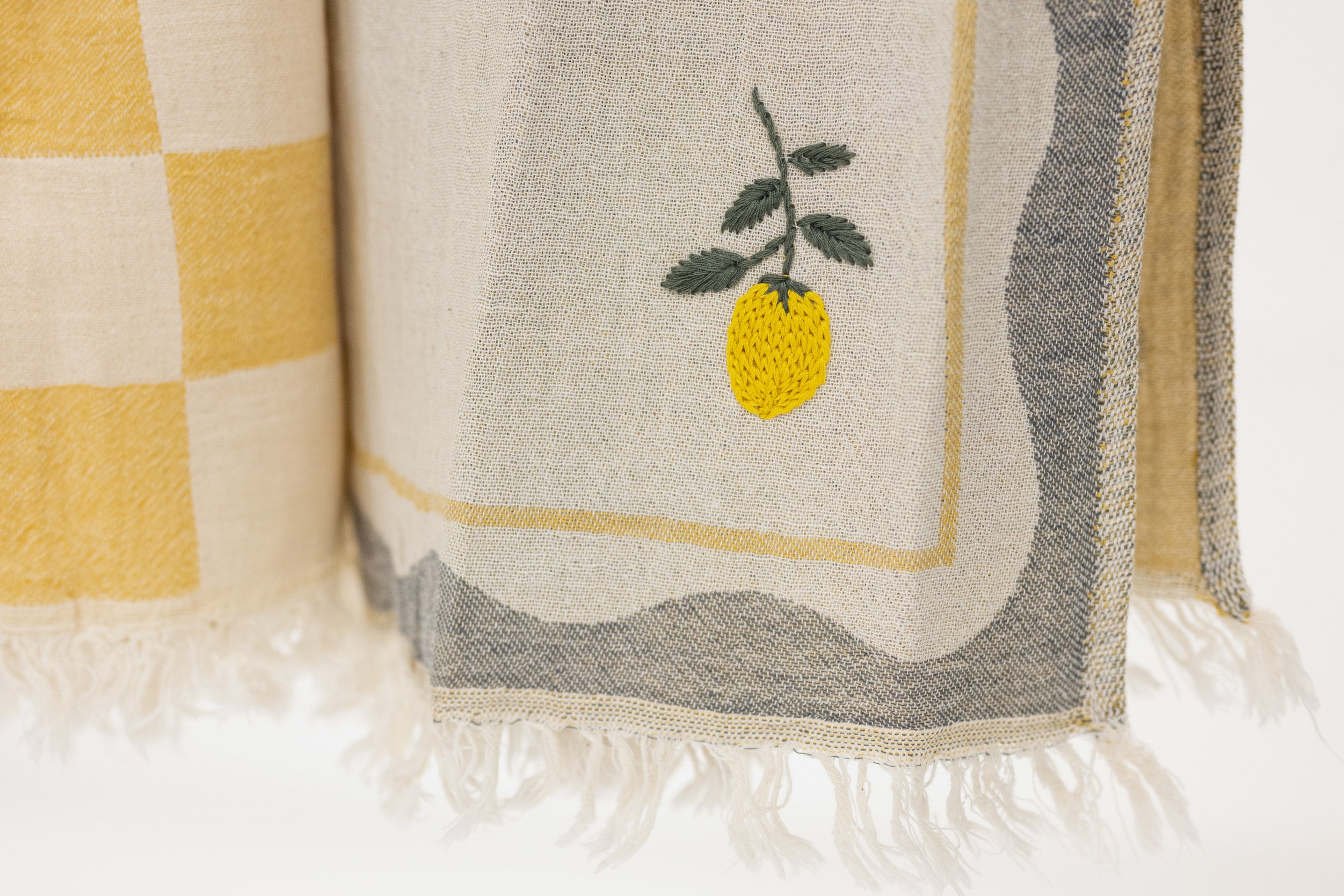 A close-up of a woven textile with fringe, featuring a yellow lemon embroidery on a cream background with gray and yellow border details.