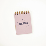 A small lilac spiral-bound notebook with the name 'ELKADER' printed in navy blue text, surrounded by small star and diamond illustrations. The notebook is photographed on a clean white background.