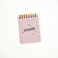 A small lilac spiral-bound notebook with the name 'ELKADER' printed in navy blue text, surrounded by small star and diamond illustrations. The notebook is photographed on a clean white background.