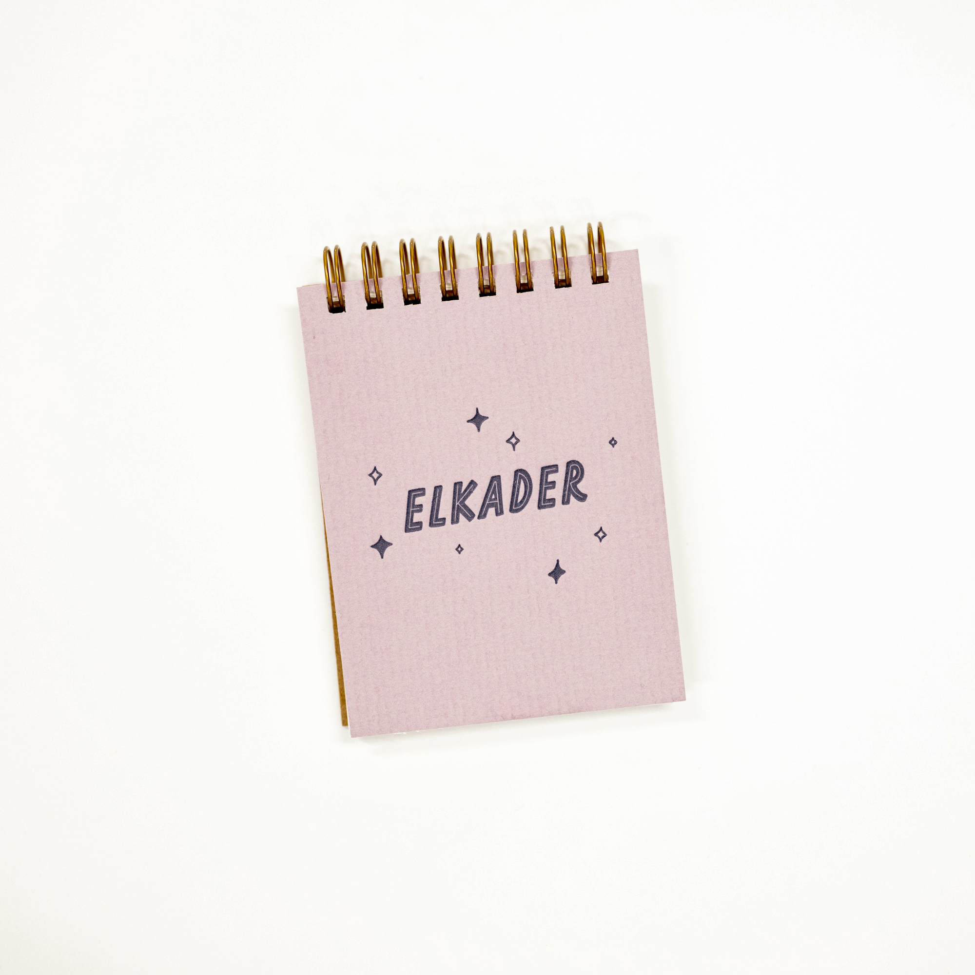 A small lilac spiral-bound notebook with the name 'ELKADER' printed in navy blue text, surrounded by small star and diamond illustrations. The notebook is photographed on a clean white background.