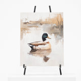 A vertical canvas painting of a mallard duck on a reflective pond, displayed on a black easel against a clean white background.