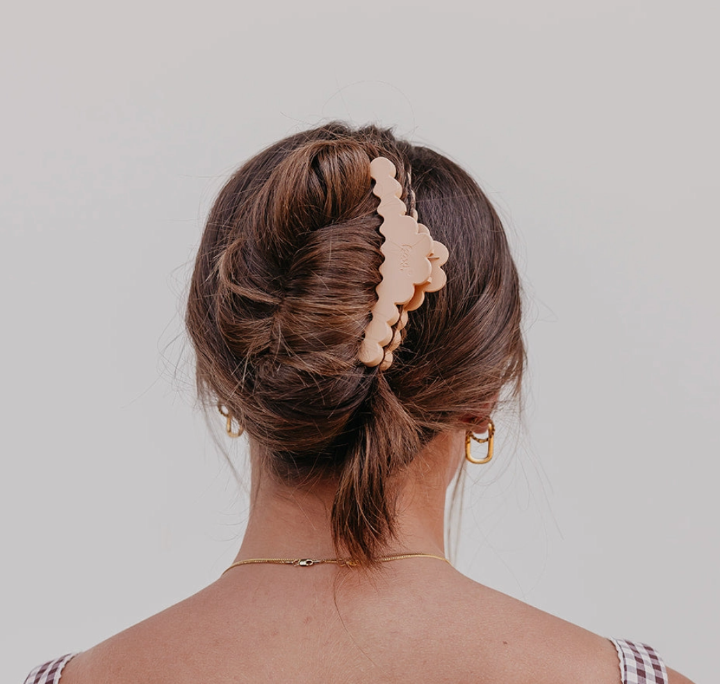 A woman viewed from the back wearing a large, scalloped-edge claw clip in a light beige or peachy-pink tone holding her brown hair in an updo. She is wearing small gold hoop earrings and a delicate gold chain necklace against a plain light gray background.