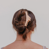 A woman viewed from the back wearing a large, scalloped-edge claw clip in a light beige or peachy-pink tone holding her brown hair in an updo. She is wearing small gold hoop earrings and a delicate gold chain necklace against a plain light gray background.