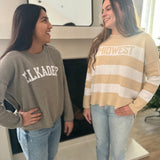 Two women standing indoors, one wearing a gray sweatshirt with 'ELKADER' and the other in a striped beige and white sweater.