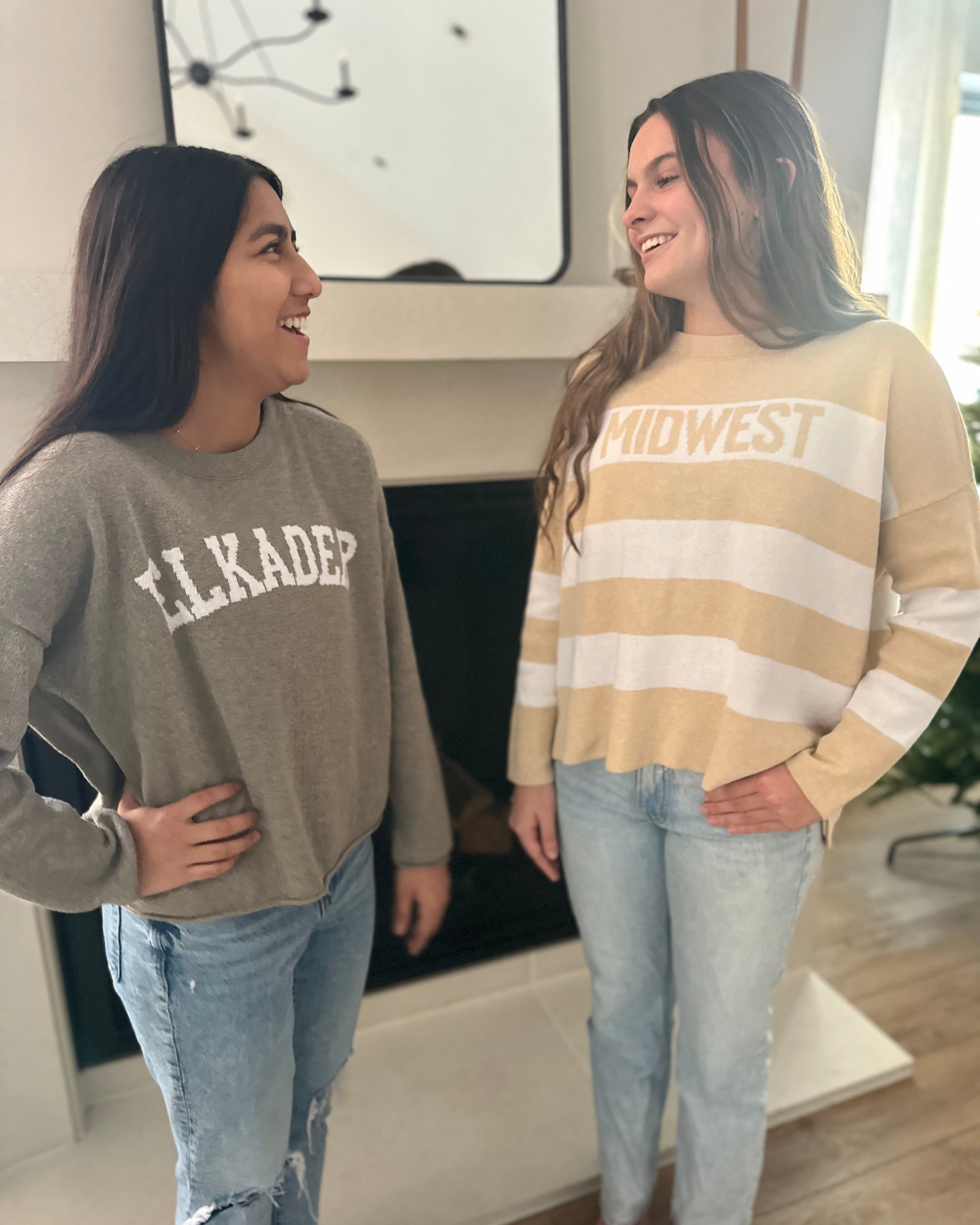 Two women standing indoors, one wearing a gray sweatshirt with 'ELKADER' and the other in a striped beige and white sweater.