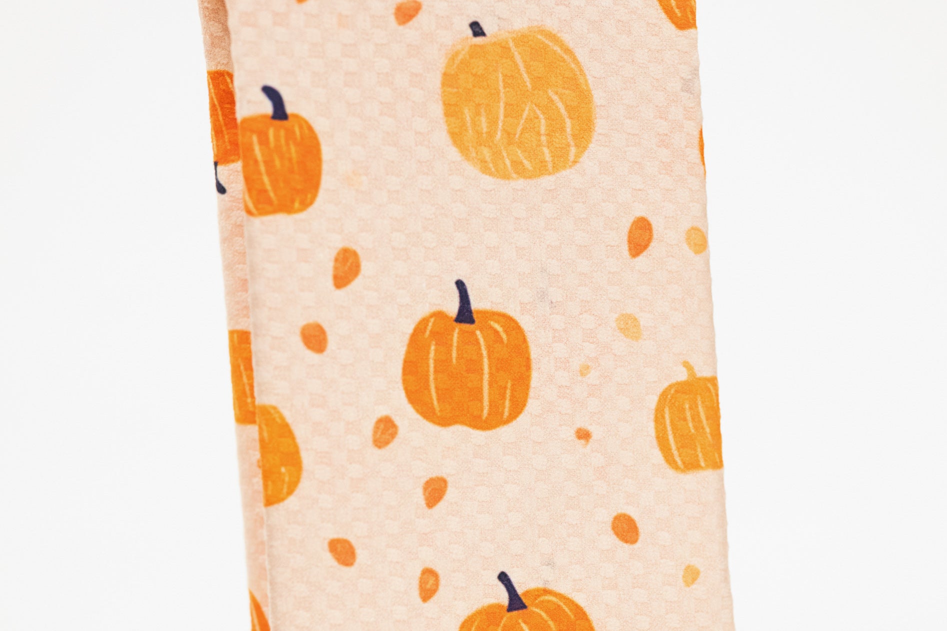 A close-up of a kitchen towel featuring a repeating pattern of orange pumpkins and small orange leaves on a light peach background.