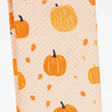 A close-up of a kitchen towel featuring a repeating pattern of orange pumpkins and small orange leaves on a light peach background.