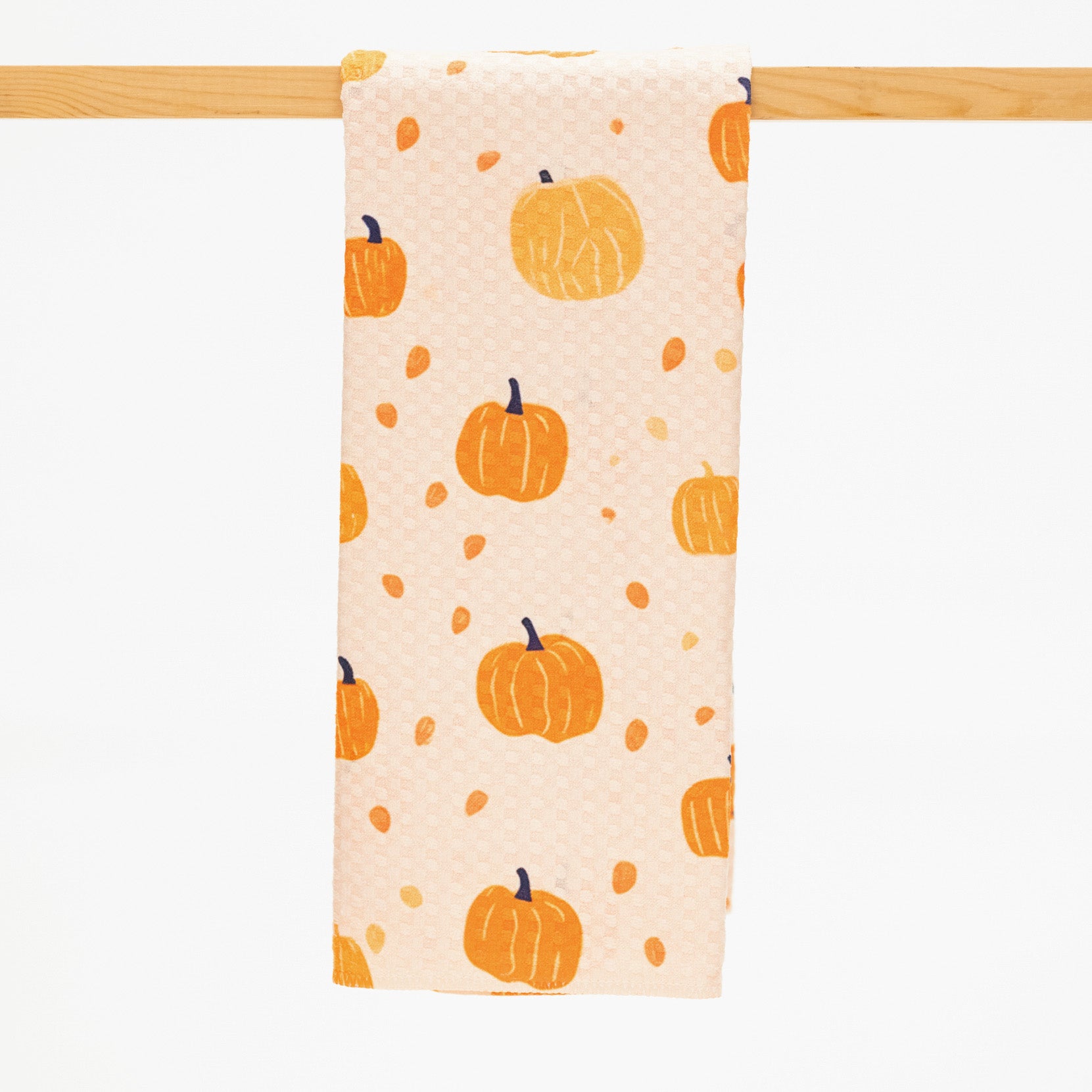 A fabric kitchen towel with an orange pumpkin and acorn pattern on a light peach background, hanging from a wooden dowel against a white background.