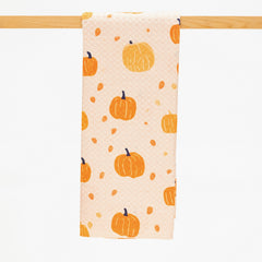 A fabric kitchen towel with an orange pumpkin and acorn pattern on a light peach background, hanging from a wooden dowel against a white background.