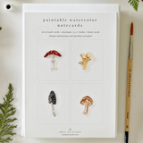 A set of paintable watercolor notecards by Emily Lee Studio, displayed on a light surface with a paintbrush and greenery accents. The notecards feature four watercolor mushroom designs on a white cardstock background.