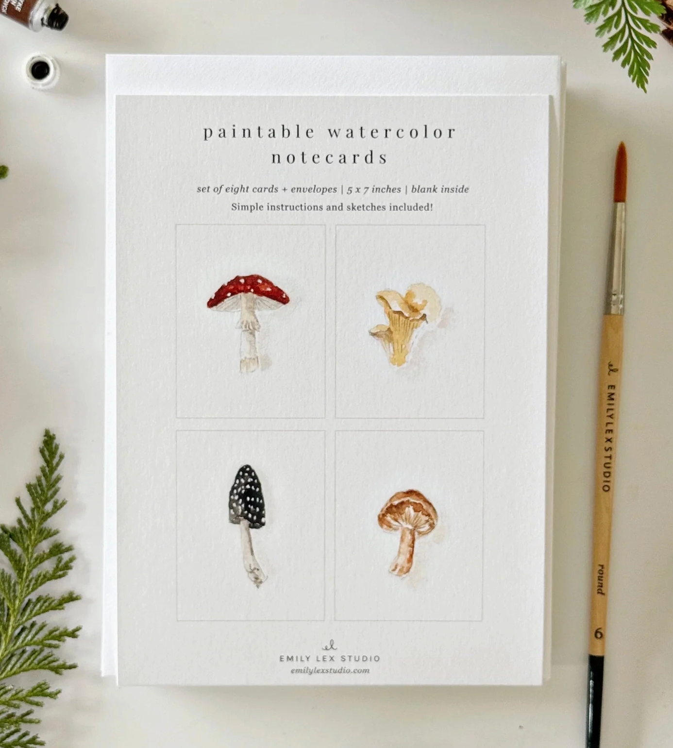 A set of paintable watercolor notecards by Emily Lee Studio, displayed on a light surface with a paintbrush and greenery accents. The notecards feature four watercolor mushroom designs on a white cardstock background.