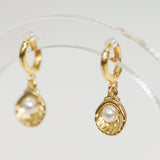 Gold shell earrings with pearl drops on a clear stand against a light background