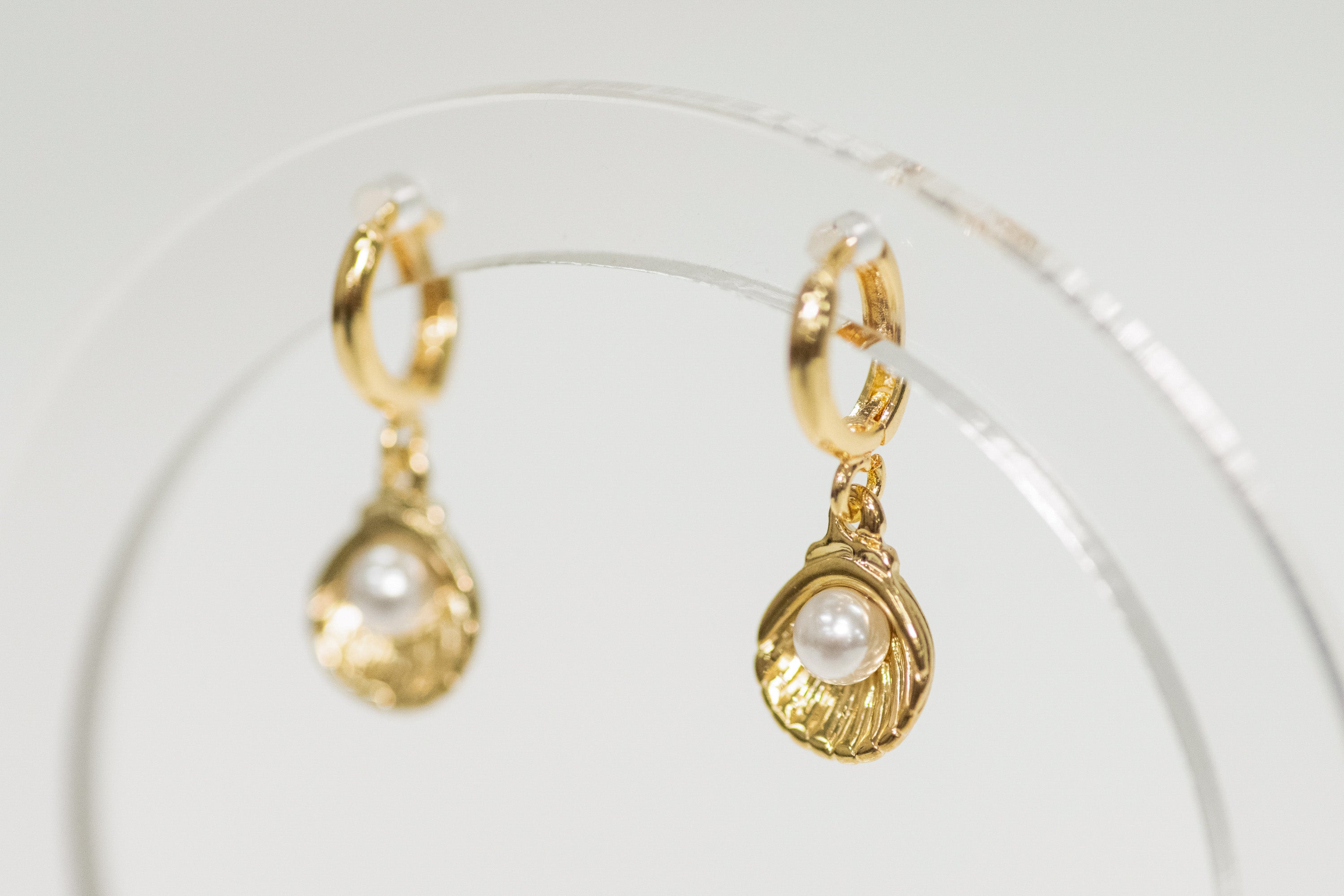Gold shell earrings with pearl drops on a clear stand against a light background